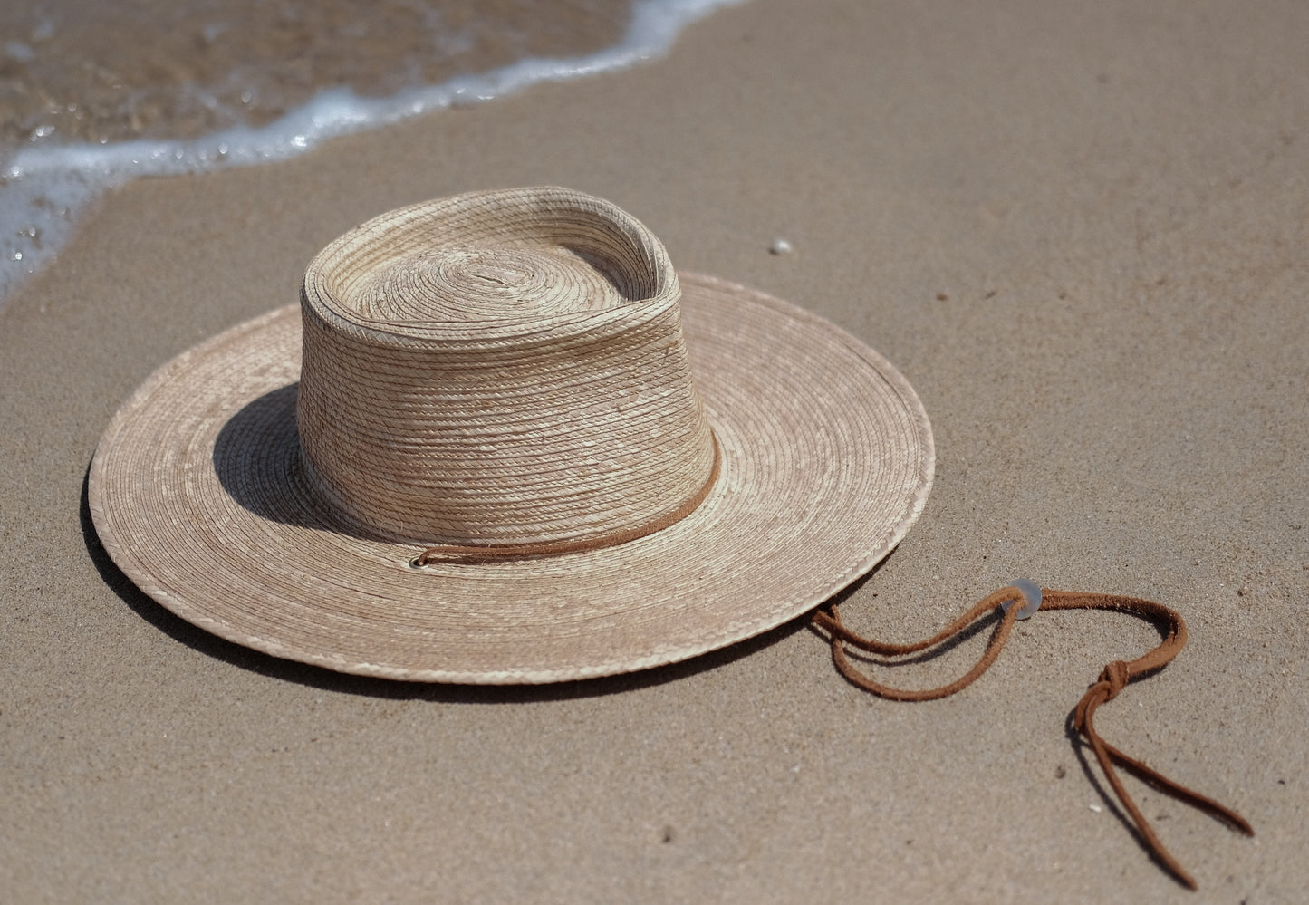 Jack Savage Wayfinder Palm straw hat on a sandy beach with ocean waves in the background